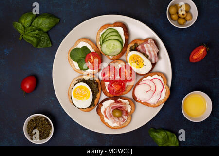 Varianten von Sandwiches mit Frischkäse, Pesto, Tomaten, Gurken, Ei, Speck, Rettiche auf grauem Hintergrund. Ansicht von oben Stockfoto