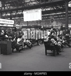 1950, historische, bahnreisende am Bahnhof St Pancras sitzen auf Bänken Zeitung lesen, während ihre Züge warten, London, England, UK. Wie in der Ferne zu sehen ist, war es immer noch die Zeit der Dampf, der sich aus den Plattformen gesehen werden kann. Die Station wurde von Midland Railway als Ihre eigenen Central London Endstation gebaut und im Jahre 1868 eröffnet, mit seinen berühmten 1-Struktur Dach oder trainshed von William Henry Barlow entworfen. Stockfoto