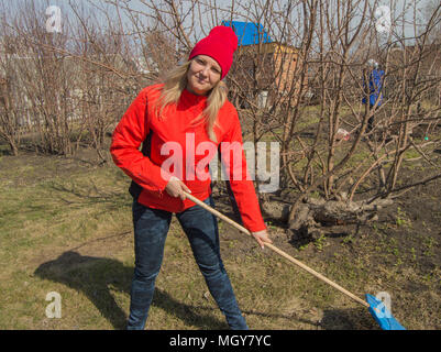Junge schöne Frau Bauer reinigt Rechen trockenes Gras, das Tragen der roten Jacke, Stiefel und Hut, vertikale Foto Stockfoto