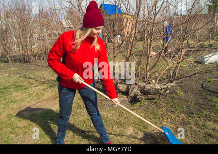 Junge schöne Frau Bauer reinigt Rechen trockenes Gras, das Tragen der roten Jacke, Stiefel und Hut Stockfoto