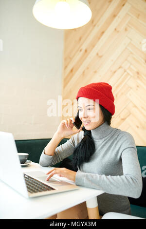 Porträt der moderne junge Frau trägt Beanie-mütze mit Laptop dabei freie Mitarbeit im Cafe, spricht von Telefon und lächelnd Stockfoto