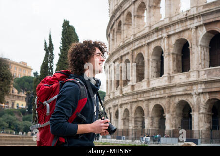 Hübscher junger Tourist Mann mit lockigem Haar mit Kamera und Rucksack, die Bilder von Kolosseum in Rom, Italien. Jungen Touristen, die Bilder von Co