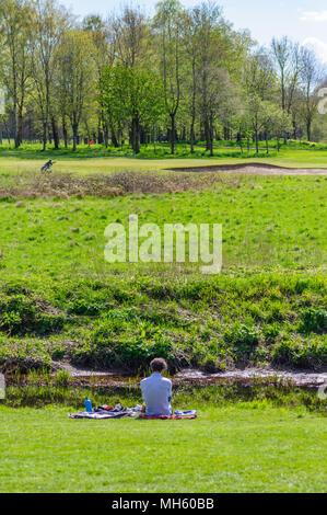 Glasgow, Schottland, Großbritannien. 30. April, 2018. UK Wetter: ein Mann sitzt auf dem Gras am Ufer der Weißen Warenkorb Wasser mit Pollok Golfkurs im Hintergrund auf einem warmen und sonnigen Tag im Pollok Country Park. Credit: Skully/Alamy leben Nachrichten Stockfoto