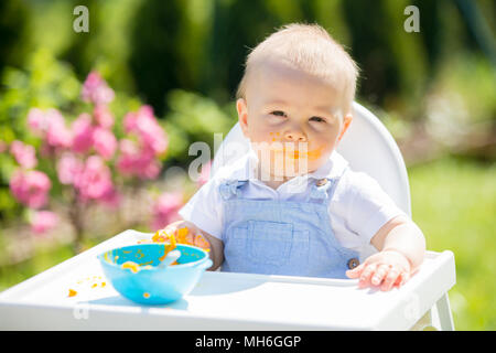 Niedliches Kleinkind mit unordentlichen Gesicht beim Mittagessen im Garten, im Baby Stuhl sitzen. Außenaufnahme. Stockfoto