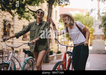 Positive und glückliche Mädchen zu Fuß auf der Straße der Stadt mit Fahrrädern. Weibliche Freunde genießen Sie einen auf der Straße mit ihren Bikes. Stockfoto