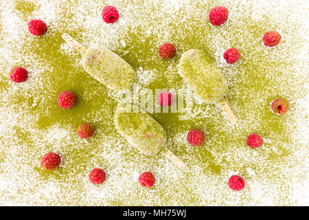 Frische Himbeeren und drei Matcha und Himbeere Eis am Stiel auf weißem Hintergrund bestäubt mit Matcha Pulver. Stockfoto