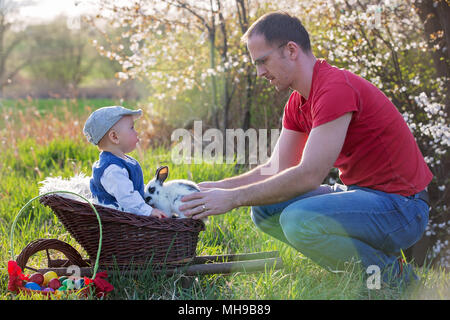 Süße kleine Baby boy in Katze in einem blühenden Garten mit kleinen Hasen und bunten Ostereier, in die Kamera lächeln Stockfoto