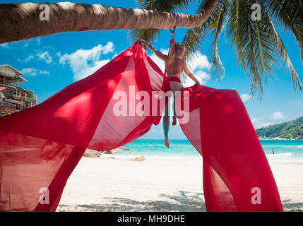 Weibliche Luftakrobat, akrobatische Tricks mit einem roten Antenne Seide an einem Strand in einer tropischen Umgebung. Stockfoto