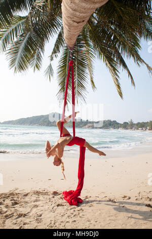 Weibliche Luftakrobat, akrobatische Tricks mit einem roten Antenne Seide an einem Strand in einer tropischen Umgebung. Stockfoto