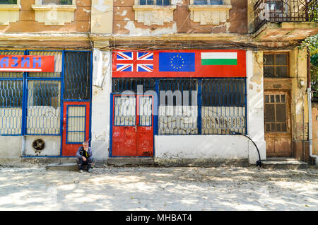 Alte Menschen sitzen, die durch eine Straße, vor einem Geschäft mit der Bulgarischen, UK und EU-Flaggen. Plovdiv, Bulgarien Stockfoto