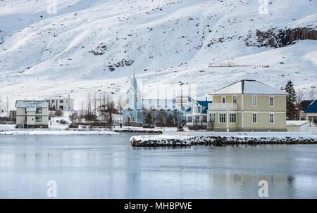 Blick auf die Blaue Kirche Seyðisfjarðarkirkja im Dorf Seyðisfjörður mit Schnee, Reflexion in den See Fjarðará Stockfoto