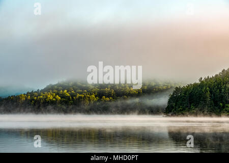 Bunte, ruhig Nebel Clearing aus bewaldeten See mit Bootshaus und Dock in der Ferne Stockfoto