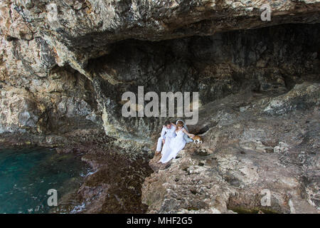 Paar Brautpaar Braut und Bräutigam Lachen und Lächeln zu einander, glücklichen und freudigen Moment. Der Mann und die Frau in der Hochzeit Kleidung sitzen auf dem Rock Hintergrund. Stockfoto