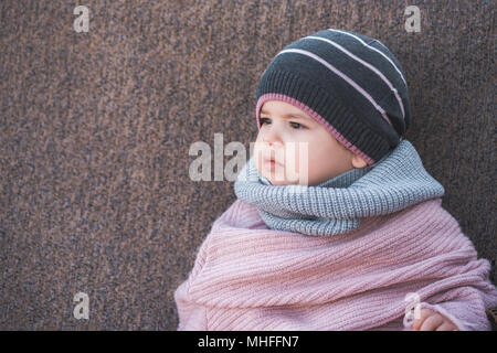 Cute Baby Mädchen mit einem warmen Winter Hut und ein bunter Schal auf braunem Hintergrund Stockfoto