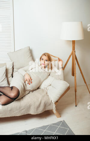 Vertikale Portrait der hübschen Blondine schwangere Frau mit roten Lippenstift das Spielen mit ihrem Haar, während auf dem Sofa in der Weißen Moderne Zimmer. Stockfoto