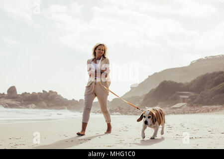 Volle Länge geschossen von reife Frau, ihren Hund zu Fuß am Strand. Ältere Weibchen auf der morgendlichen Spaziergang mit ihrem Hund. Stockfoto