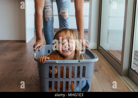 Schöne Mädchen in einer Waschmaschine Korb von ihrer Mutter geschoben zu sitzen. Süße kleine Mädchen und ihre Mutter während des Spielens Wäscherei zu Hause zu tun. Stockfoto