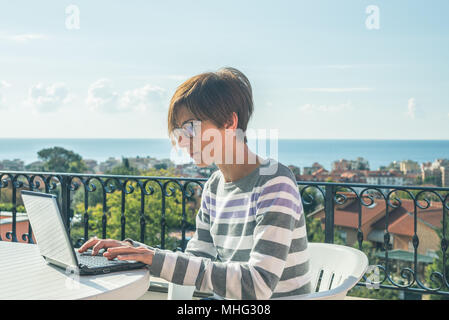 Frau mit Brille und legere Kleidung am Laptop im Freien auf der Terrasse. Schönen Hintergrund von grünen Bergen und blauem Himmel in einem hellen, sonnigen Morgen Stockfoto