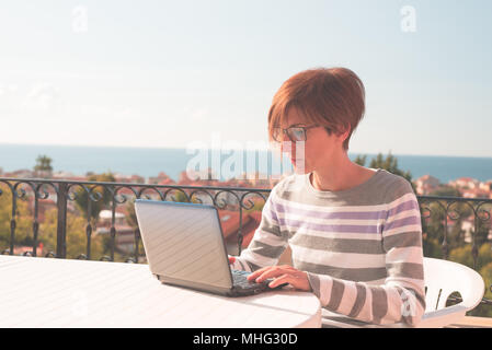 Frau mit Brille und legere Kleidung am Laptop im Freien auf der Terrasse. Schönen Hintergrund von grünen Bergen und blauem Himmel in einem hellen, sonnigen Morgen Stockfoto