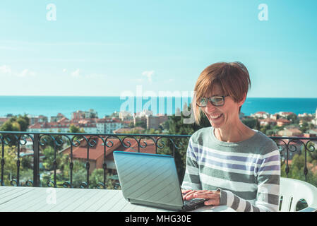 Frau mit Brille und legere Kleidung am Laptop im Freien auf der Terrasse. Schönen Hintergrund von grünen Bergen und blauem Himmel in einem hellen, sonnigen Morgen Stockfoto