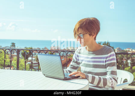 Frau mit Brille und legere Kleidung am Laptop im Freien auf der Terrasse. Schönen Hintergrund von grünen Bergen und blauem Himmel in einem hellen, sonnigen Morgen Stockfoto