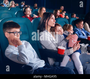 Die Zuschauer sitzen in bequemen Stühlen und beobachten Film im Kino. Junge schauen mit Interesse am Bildschirm mit unerschütterlichem Augen. Kleine hübsche Mädchen mit geschlossenen Augen sitzen auf ihrer Mama runden. Stockfoto