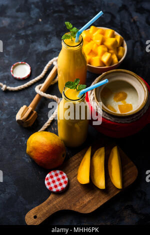 Mango Lassi oder Smoothie in großen Glas oder kleine Flaschen mit Quark, Obst Stücke schneiden und Mixer. Moody Hintergrund, selektiver Fokus Stockfoto