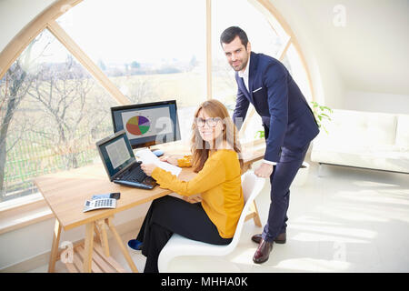 Lächelnd reife Geschäftsfrau, Sitzen im Büro, während ihre junge Kollegin stand neben ihr und gemeinsam mit dem Finanzbericht. Teamarbeit Stockfoto