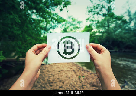 Frau Hände halten ein Blatt Papier mit Bitcoin Symbol in der Mitte der Natur mit einem Fluss in der Nähe der Wald fließt. Eco crypto und digitale Wechselstu Stockfoto