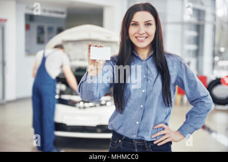 Eine schöne Frau hält eine Visitenkarte des Auto Service Center. Prüft der Mechaniker das Auto unter der Haube im Hintergrund Stockfoto