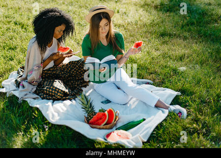 Zwei junge hübsche afrikanische und kaukasischen girl friends essen die Wassermelone auf einem Picknick. Die brünette ist Chatten und Surfen im Internet über das Mobiltelefon, während ihre blonde Freundin das Buch zu lesen. Stockfoto