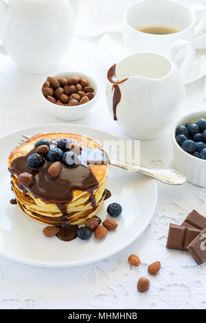 Das traditionelle Frühstück Konzept. Stapel Pfannkuchen mit Schokoladensauce, Blaubeeren und die Muttern an der weißen Tischdecke. Selektiver Fokus Stockfoto