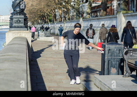 Plogging UK. Frau Joggen entlang der Southbank durch den Fluss in London, herauf und Wegwerfen von Müll in einem Abfallbehälter. Stockfoto
