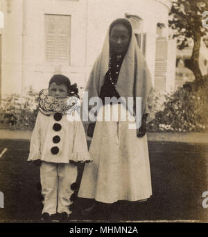 Ein kleiner Junge, gekleidet in einen Clown Kostüm für eine Party. Er scheint durch seine Krankenschwester oder Ayah - das Foto sieht auf dem indischen Sub - Kontinent ergriffen werden begleitet werden. Datum: 1920 s Stockfoto