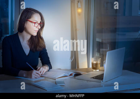 Frau mit Brille sitzt in einem hölzernen Tisch bei Nacht, Studieren und Schreiben ihren Laptop suchen Sie bei einem Kaffee. Stockfoto
