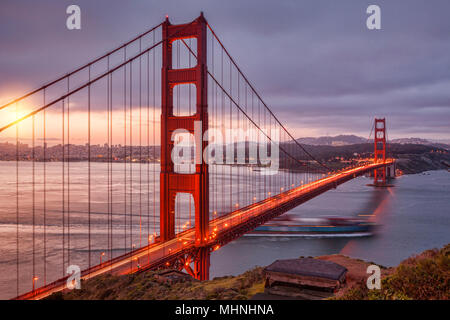 Die Golden Gate Bridge, San Francisco, von der Batterie Spencer in der Dämmerung, mit Verkehr auf der Brücke und einem Container schiff aus dem Schacht heraus. Stockfoto