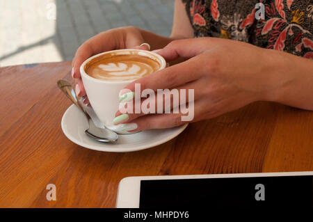 Die junge Dame, mit wunderschönen Maniküre Holding eine Tasse Cappuccino und Latte Kunst auf hölzernen Tisch in einem Cafe. Stockfoto