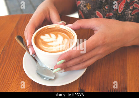 Die junge Dame, mit wunderschönen Maniküre Holding eine Tasse Cappuccino und Latte Kunst auf hölzernen Tisch in einem Cafe. Stockfoto