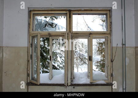 Alte Fenster in verlassenen Haus mit Glasscherben und schneebedeckten Fensterrahmen umgeben mit verfallenen Mauern und Schnee Bäume im Hintergrund Stockfoto