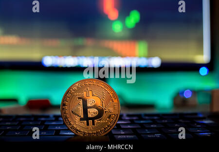 Physische Bitcoin auf Laptop mit Charts auf dem Bildschirm platziert, dunkle themed Stockfoto