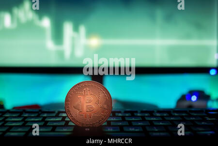 Physische Bitcoin auf Laptop mit Charts auf dem Bildschirm platziert, dunkle themed Stockfoto