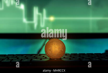 Physische Bitcoin auf Laptop mit Charts auf dem Bildschirm platziert, dunkle themed Stockfoto