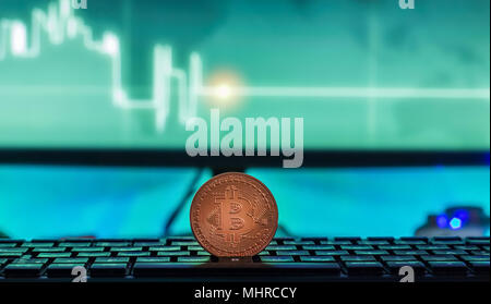 Physische Bitcoin auf Laptop mit Charts auf dem Bildschirm platziert, dunkle themed Stockfoto