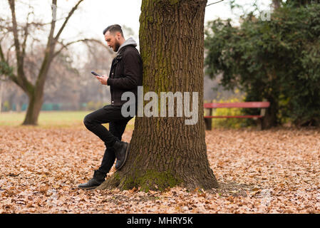 Ein hübscher junger hipster Mann lehnte sich an einen Baum und sein Smartphone Kontrolle in den Park. Stockfoto