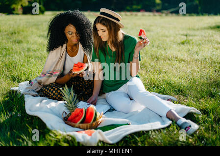 Timespending von zwei multi-ethnischen Freundinnen. Ziemlich afrikanische Mädchen über die mobile und die Wassermelone zu essen, während Ihre attraktive Blondine Freund ist lesen Buch und das Essen von Früchten. Picknick Thema plaudern ist. Stockfoto