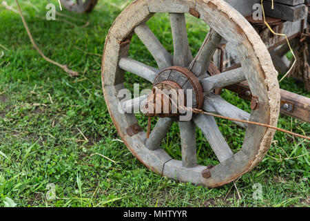 Holz- Rad eines alten Warenkorb auf einem Boden mit grünem Gras bedeckt. Rostiges Metall, verwittertes Holz Stockfoto