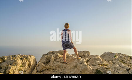 Blonde Frau mittleren Alters in blauem Kleid grüßt die Sonne Dawn in die Ferne sieht und das Leben zu genießen, im Sommer Konzept Stockfoto
