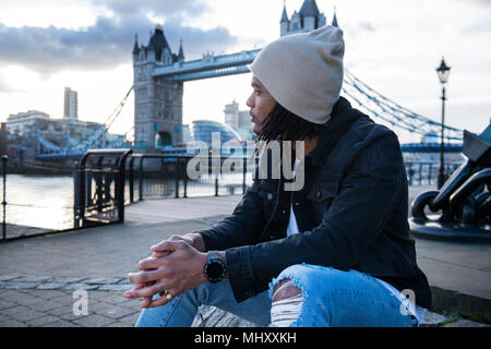 Junge Mann sitzt im Freien, nachdenklich, Ausdrücke, die Tower Bridge im Hintergrund, London, England, Großbritannien Stockfoto