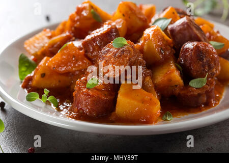 Kartoffeln Eintopf mit Schweinefleisch Wurstscheiben und Tomatensauce auf einem weißen Teller mit Oregano Blätter Stockfoto