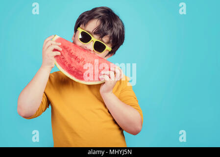 Glückliches Kind mit Sonnenbrille isst Wassermelone auf blauem Hintergrund glückliches Kind mit Sonnenbrille isst Wassermelone auf blauem Hintergrund Stockfoto